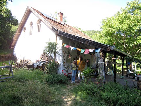 Istvan Sky mit Svetlana auf der Veranda vor seinem Haus, umgeben von grünem Gras. Rechts der von István gepflanzte Mandelbaum. Szentbékkállá.