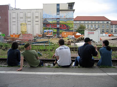Участники проекта Graffiti Workshop 2004, сидя на перроне перед  ж/д путями смотрят на стену гостиницы InterCityHotel, Эрфурт.