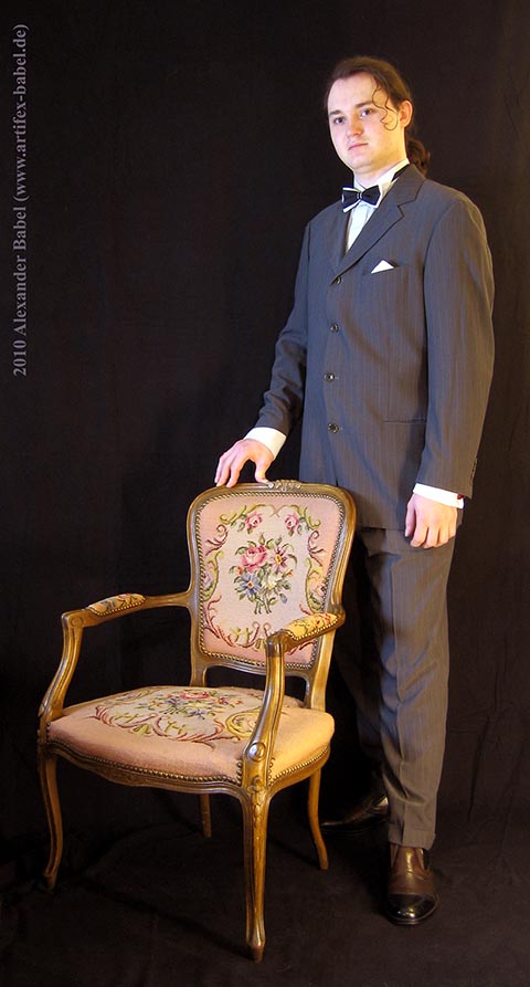 Александр Бабель крупным планом в полный рост в анфас. В костюме, с бабочкой. Рядом антикварный стул.