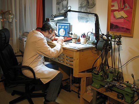 Александр Бабель работает за столом часовщика, рядом на подставке токарный станок Schaublin 70, на стенах авторские рисунки.